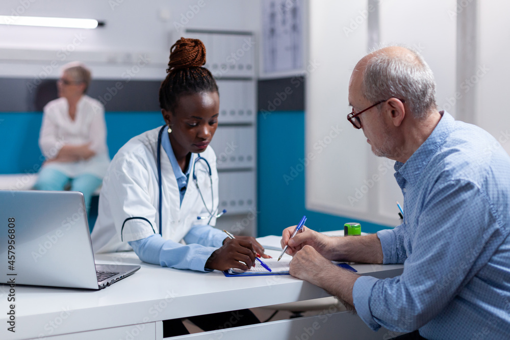 Obraz premium Black woman with medic profession looking at documents with elderly patient sitting at white desk. African american doctor and old man with disease writing on paper files for checkup