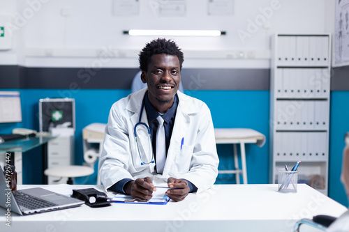 Portrait of african american doctor sitting at desk in medical cabinet with laptop computer and equipment used for healthcare examination. Black medic with office uniform and stethoscope