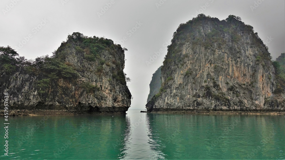 There is a narrow passage between the islands in Halong Bay. There is scant vegetation on the steep rocky slopes. Foggy sky. Reflection on the water surface. Vietnam There is a narrow passage between the islands in Halong Bay. There is scant vegetation on the steep rocky slopes. Foggy sky. Reflection on the water surface. Vietnam