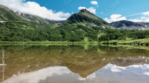 A picturesque mountain range on the shore of a lake. On the slopes there is green vegetation and areas of melted snow. A mirror reflection on calm water. Clouds in a blue sky. Kamchatka. Vachkazhets