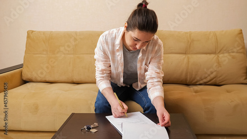 Young woman holding pen reads apartment purchase contract before signing at small coffee table with keys sitting on comfortable sofa in room