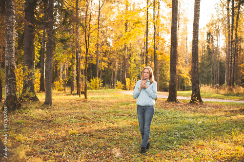 A beautiful happy blonde woman walks through the autumn park with a cup of warming drink in her hands. Walk on a sunny autumn evening. Healthy lifestyle.