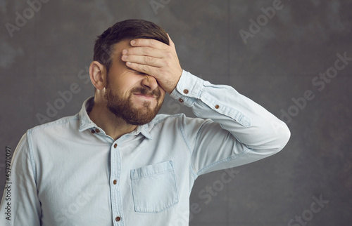 Businessman covers face with hand. Guy facepalms feeling ashamed of terrible mistake or poor memory. Annoyed guilty cringing business manager employee closes face with palm isolated on grey background photo