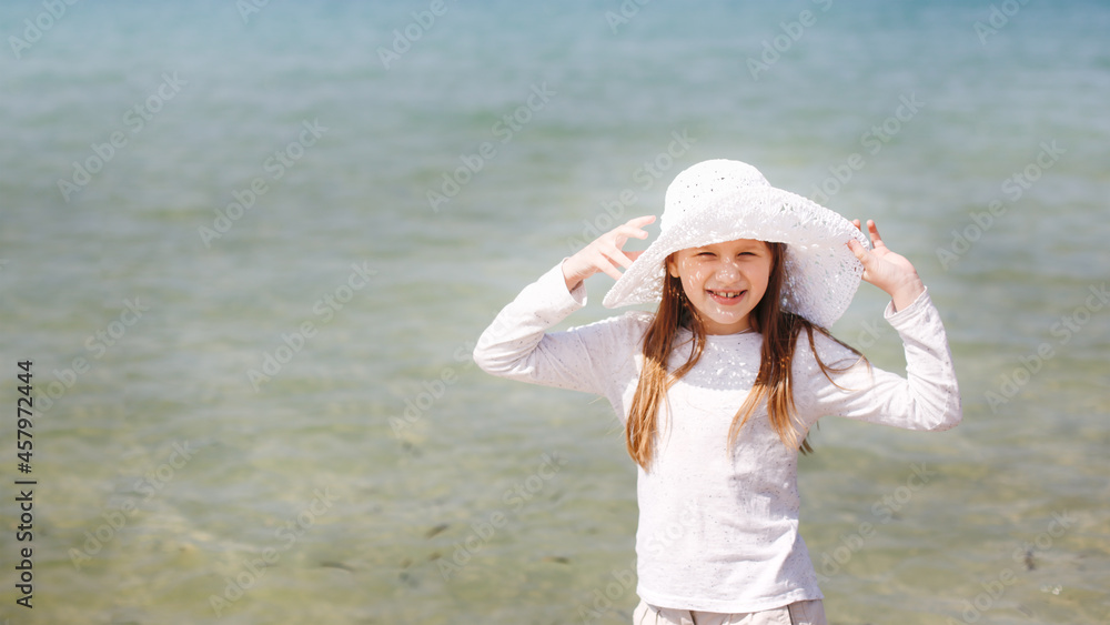 Cute caucasian child girl with long hair by sea. A child in hat and long sleeve with shorts, protection from sun, first spring sea, the beginning of sea season. Childhood and freedom, joy from sea