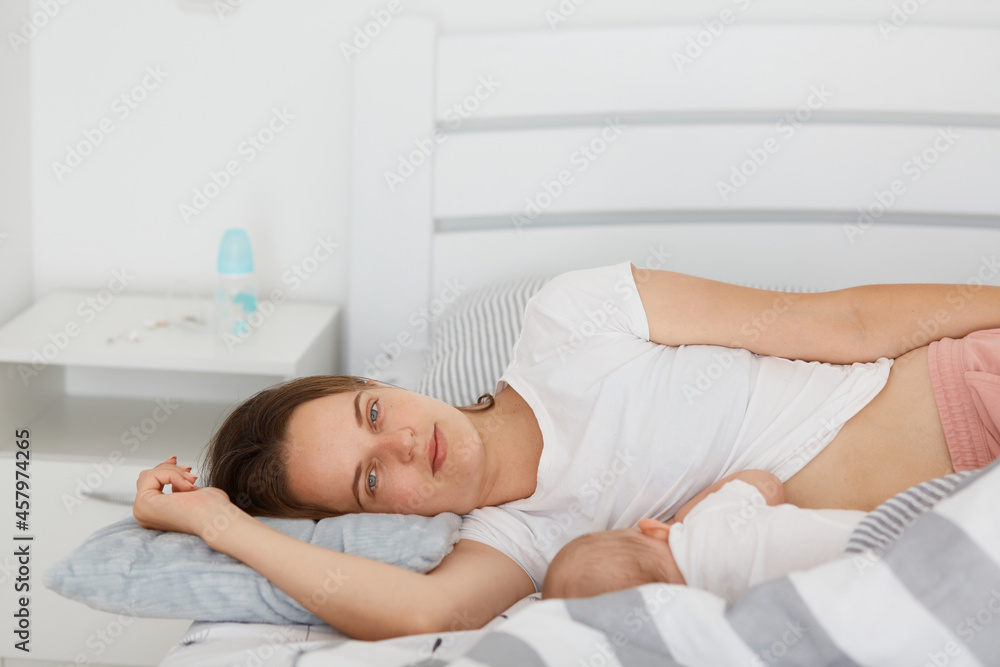 Portrait of young mother lying at her baby sleeping in bed, woman looking at camera with happy but tired look, having exhausted facial expression, posing light room. Portrait of young mother lying at her baby sleeping in bed, woman looking at camera with happy but tired look, having exhausted facial expression, posing light room.