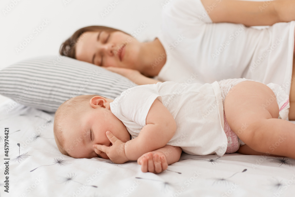 Side view of of young adult mother and her baby sleeping together lying on bed, posing at home in light room, mommy can have a rest while daughter sleeps. Side view of of young adult mother and her baby sleeping together lying on bed, posing at home in light room, mommy can have a rest while daughter sleeps.