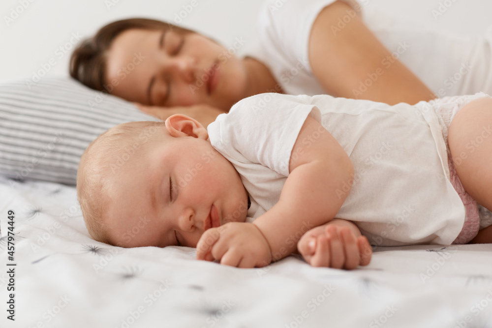 Portrait of cute charming baby girl wearing white clothing lying and sleeping near her lovely mother on bed, having nap, resting at home near mommy with closed eyes. Portrait of cute charming baby girl wearing white clothing lying and sleeping near her lovely mother on bed, having nap, resting at home near mommy with closed eyes.