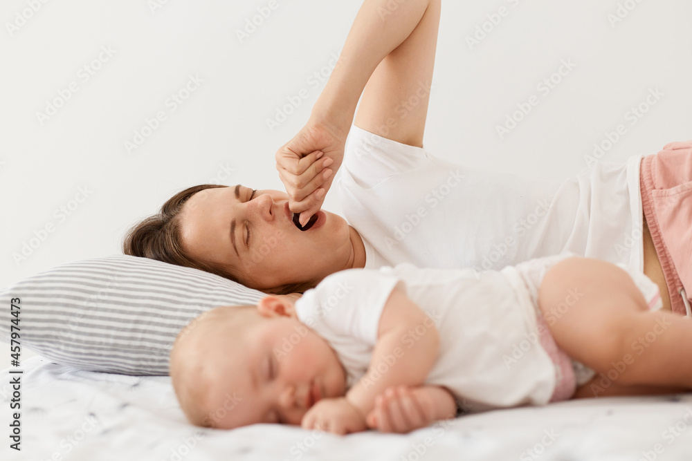 Portrait of tired exhausted mother lying on bed and yawning, covering open mouth with fist, wakes up early while her baby girl sleeping, posing at home in light room. Portrait of tired exhausted mother lying on bed and yawning, covering open mouth with fist, wakes up early while her baby girl sleeping, posing at home in light room.
