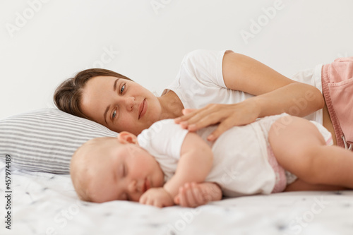 Happy pleased young dark haired mother and her baby lying together on bed, mommy hugging her charming daughter, people wearing white clothing, motherhood and childhood.
