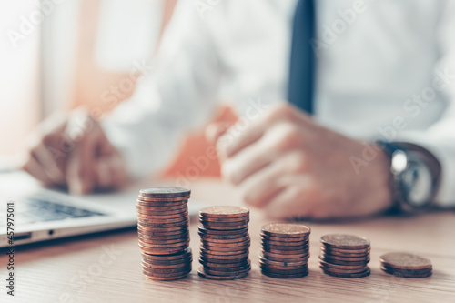 Coin stack in business office, businessman working on laptop computer in background
