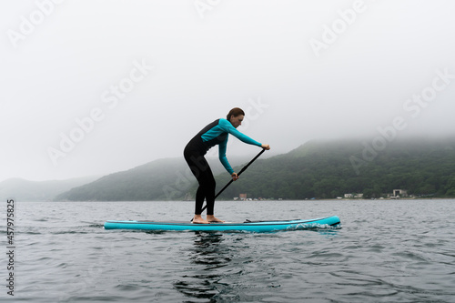 A woman in a wetsuit paddle a paddleboard with an oar on the sea waves in cloudy weather. © Aboltin