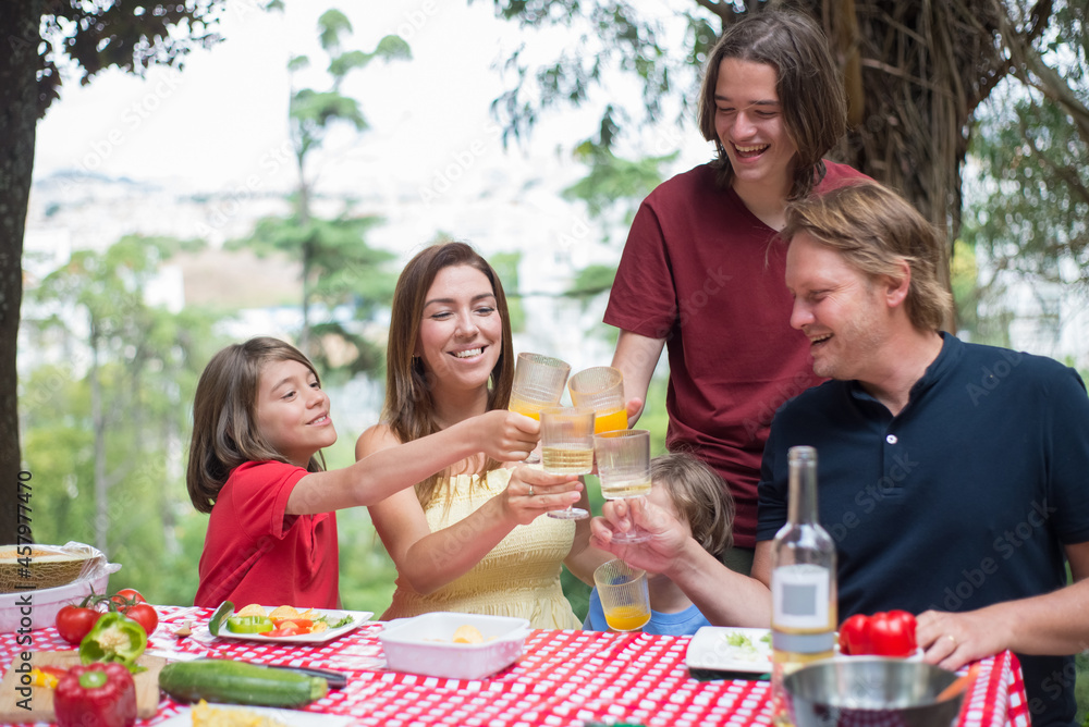 Portrait of happy family with children clinking glasses. Mid adult parents sitting around table with sons clinking glasses with yellow beverages. Family time, outdoor activity, picnic concept Portrait of happy family with children clinking glasses. Mid adult parents sitting around table with sons clinking glasses with yellow beverages. Family time, outdoor activity, picnic concept