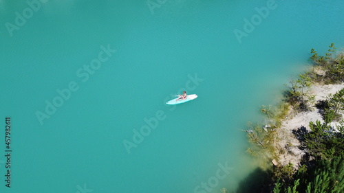 Fototapeta Naklejka Na Ścianę i Meble -  A girl in a dress floats on a glanders board on a pond with bright turquoise water. Warm summer day for travel. Top view from a quadcopter. Aerial photography