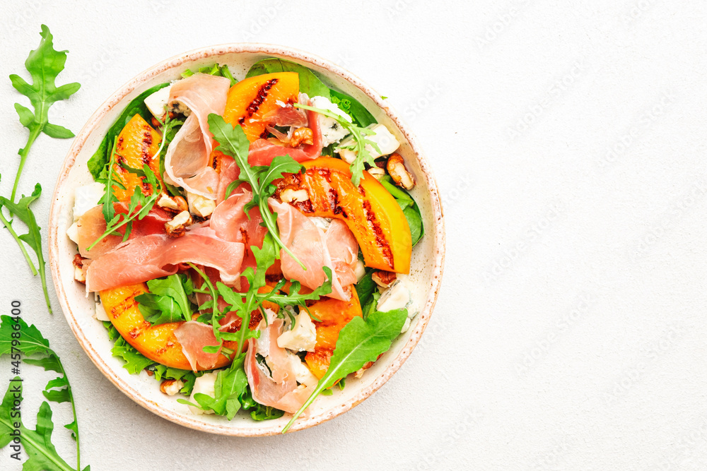 Salad bowl with grilled peach, prosciutto, goat cheese, walnuts and arugula on white background, top view, copy space Salad bowl with grilled peach, prosciutto, goat cheese, walnuts and arugula on white background, top view, copy space
