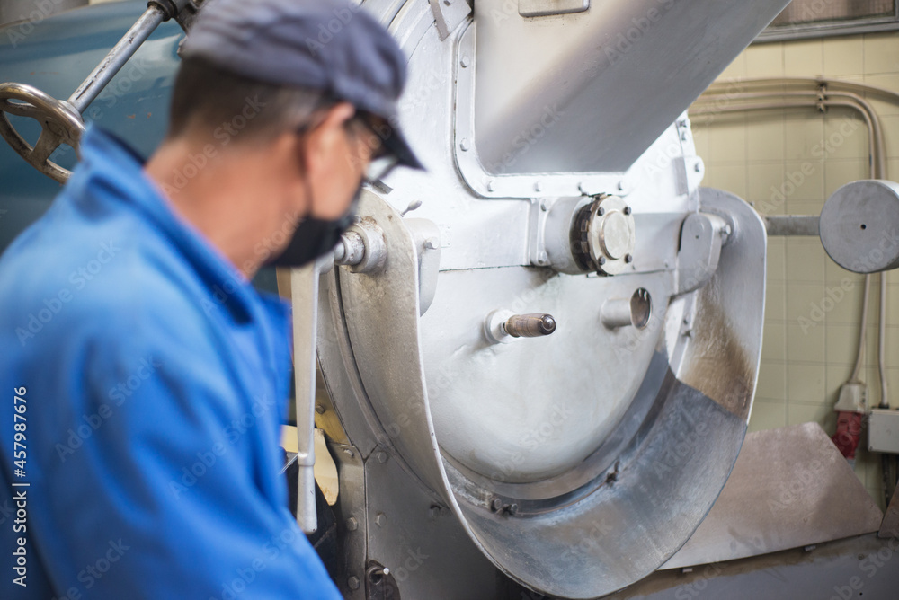 Obraz premium Worker looking at roasting machine. Partial view of coffee roaster working on roasting equipment. Man in mask and uniform Working with machinery appliance. Job, factory, maintenance concept