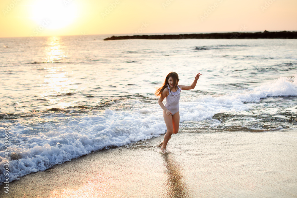 Cute caucasian girl in a striped swimsuit with long hair runs seashore on a deserted beach at a resort, evening and sunset