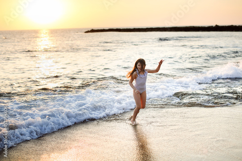 Cute caucasian girl in a striped swimsuit with long hair runs seashore on a deserted beach at a resort, evening and sunset © natalialeb
