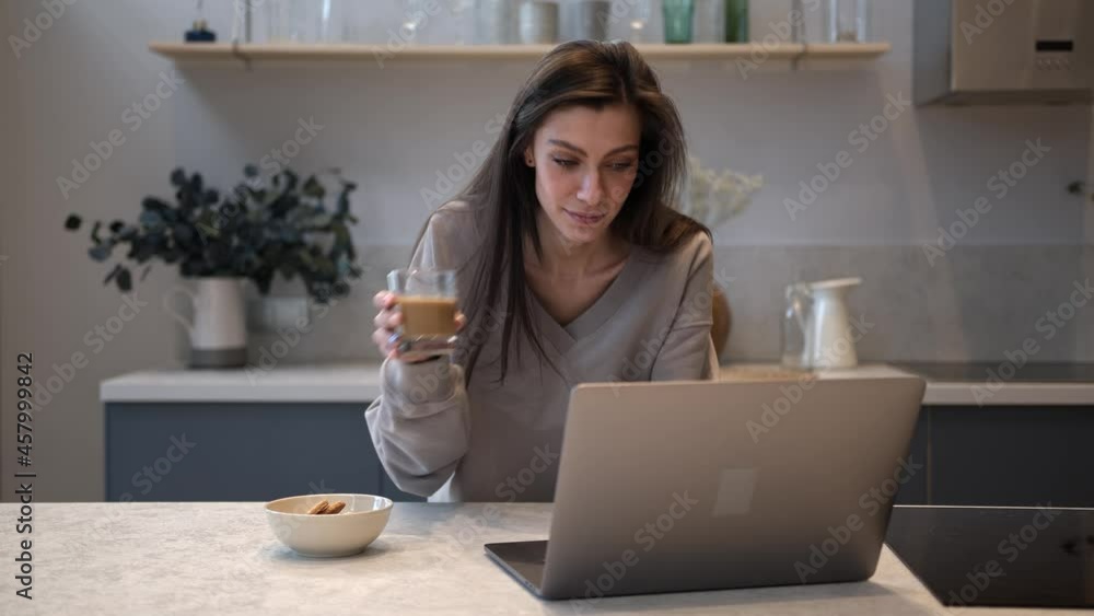 Brunette woman working in kitchen, drinking glass of a hot coffee and plate on the table. Talking on a video call in laptop at modern room. Concept of breakfast in the morning, remote work Brunette woman working in kitchen, drinking glass of a hot coffee and plate on the table. Talking on a video call in laptop at modern room. Concept of breakfast in the morning, remote work