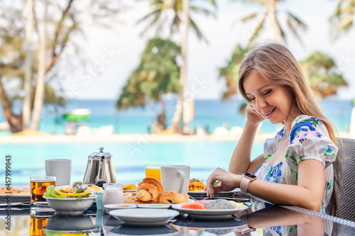 Beautiful Young Smiling Woman with Long Hair Eat a Delicious Breakfast at Resort Restaurant by the Pool with Palm Trees and Turquoise Sea on Background. Vacations or Holidays in Tropical Hotel. © TravelMedia