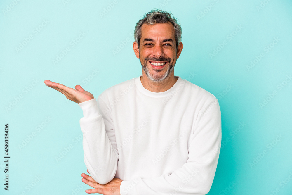 Middle age caucasian man isolated on blue background showing a copy space on a palm and holding another hand on waist. Middle age caucasian man isolated on blue background showing a copy space on a palm and holding another hand on waist.