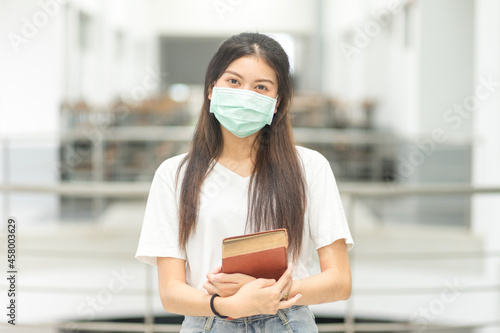 Front view portrait of a young Asian adolescent woman college student in relaxed casual wear back to school with medical face mask holds books in campus building