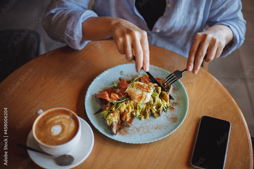 middle-aged woman in a blue shirt eats scrambled eggs and cappuccino for breakfast in a cafe