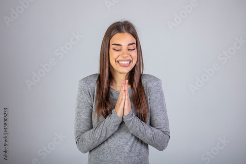 Image of happy young woman wearing sweater smiling and praying with palms together isolated over gray background. Woman asking for forgiveness smiling confident. © Dragana Gordic