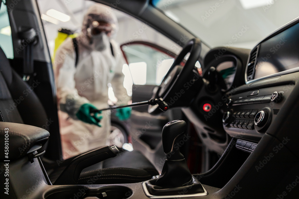 Naklejka premium COVID 19 situation warning. A man in a protective white uniform and a sprayer prevents the spread of the infection inside of a car, showroom. Stay responsible, wear the mask, coronavirus, virus