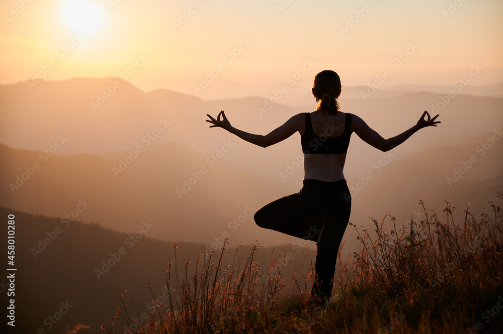 Back view of young woman performing yoga pose on grassy hill with orange sky on background. Fit woman standing on one leg and doing Gyan mudra hand gesture while doing yoga exercise outdoors.