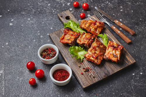 Grilled baked pork ribs with spices and vegetables on wooden cutting board on dark background. American food concept.