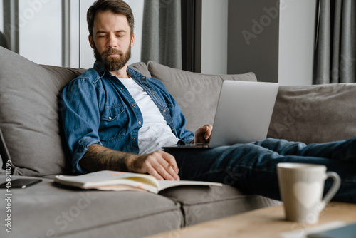 Bearded man writing down notes while working with laptop on sofa