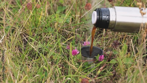 The tourist poured hot coffee from a thermos into the lid of the thermos and put it on the grass. Travel accessories. Morning drink outdoors. Travel videos.