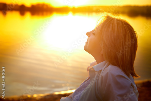 Portrait of middle-aged woman near lake or river during beautiful sunset with sun above the horizon and yellow reflection in the water