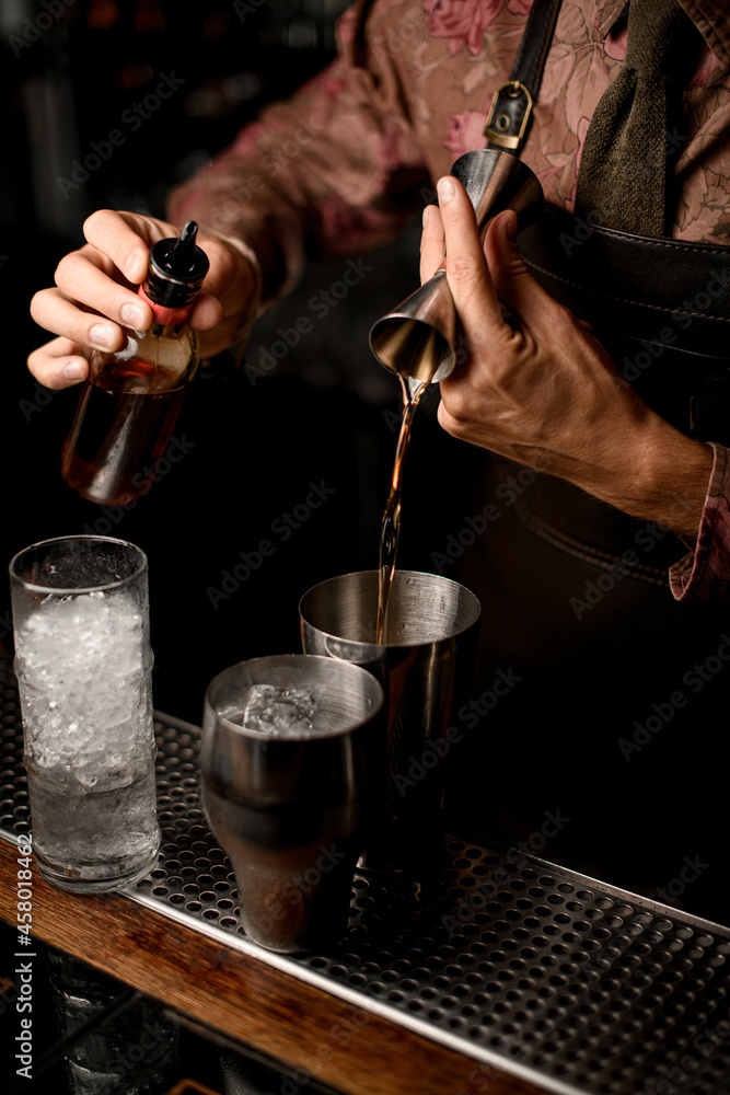 hand of bartender holds bottle with dispenser and other hand pours drink from jigger into shaker glass