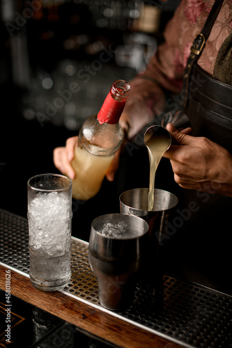 hand of bartender holds bottle and other hand pours drink from jigger into shaker glass