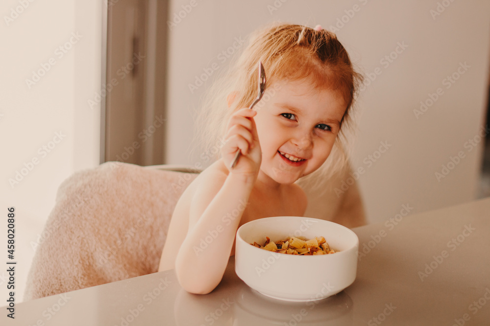 Happy toddler girl sitting at table in kitchen and eating fresh cooked meal from white plates. Tasty lunch at cozy home. Happy toddler girl sitting at table in kitchen and eating fresh cooked meal from white plates. Tasty lunch at cozy home.
