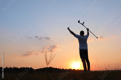 Man raising elbow crutch up to sky outdoors at sunset, back view and space for text. Healing miracle © New Africa