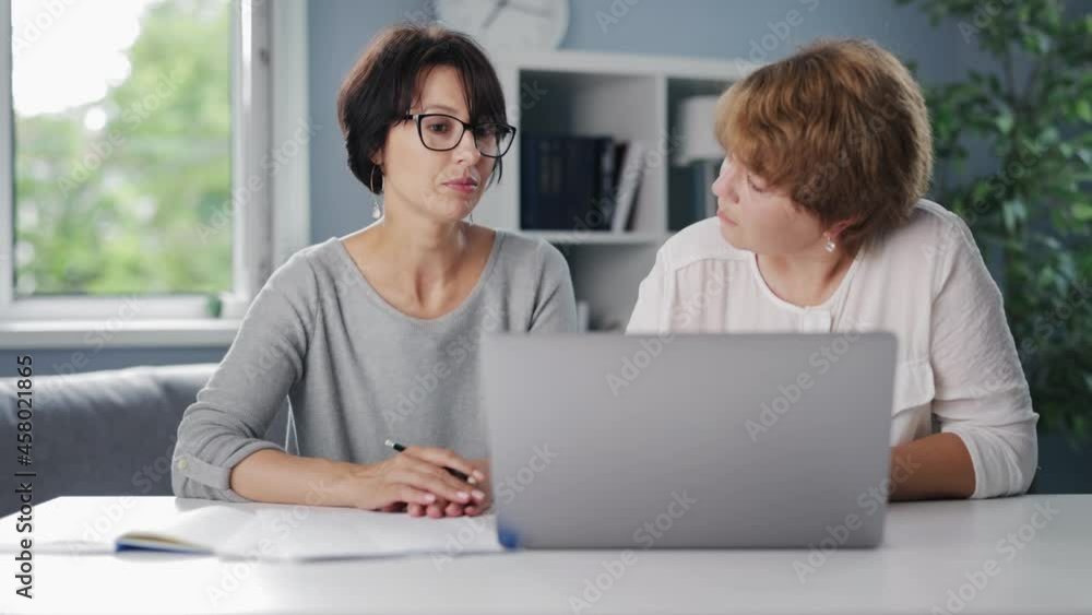 Two female business partners sitting together at table with laptop and documents and discussing common project. Concept of people, technology and cooperation. Two female business partners sitting together at table with laptop and documents and discussing common project. Concept of people, technology and cooperation.