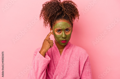 Young african american woman wearing a bathrobe and facial mask isolated on pink background pointing temple with finger, thinking, focused on a task.