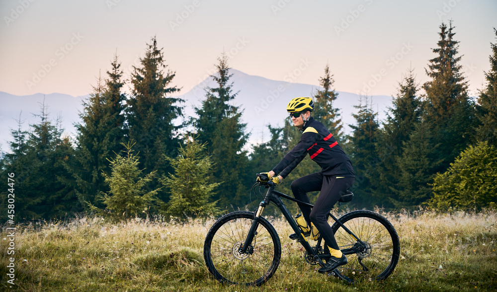 Side view of cyclist in cycling suit riding bike with coniferous trees and hills on background. Man bicyclist wearing safety helmet and glasses while enjoying bicycle ride in mountains in the morning. Side view of cyclist in cycling suit riding bike with coniferous trees and hills on background. Man bicyclist wearing safety helmet and glasses while enjoying bicycle ride in mountains in the morning.