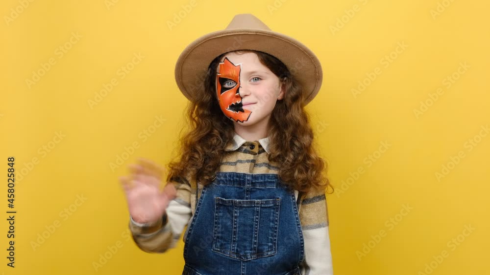 Portrait of scary bright alive fun girl kid with halloween makeup, dressed in hat and shirt, waving her hand in hello gesture standing isolated over yellow studio background. Party holiday concept Portrait of scary bright alive fun girl kid with halloween makeup, dressed in hat and shirt, waving her hand in hello gesture standing isolated over yellow studio background. Party holiday concept