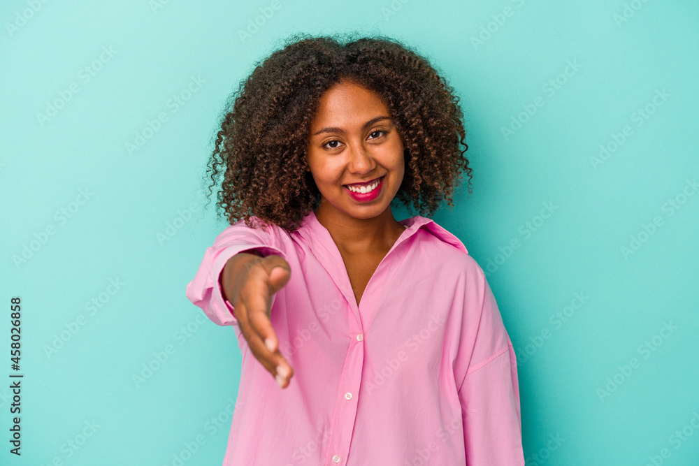 Fototapeta premium Young african american woman with curly hair isolated on blue background stretching hand at camera in greeting gesture.