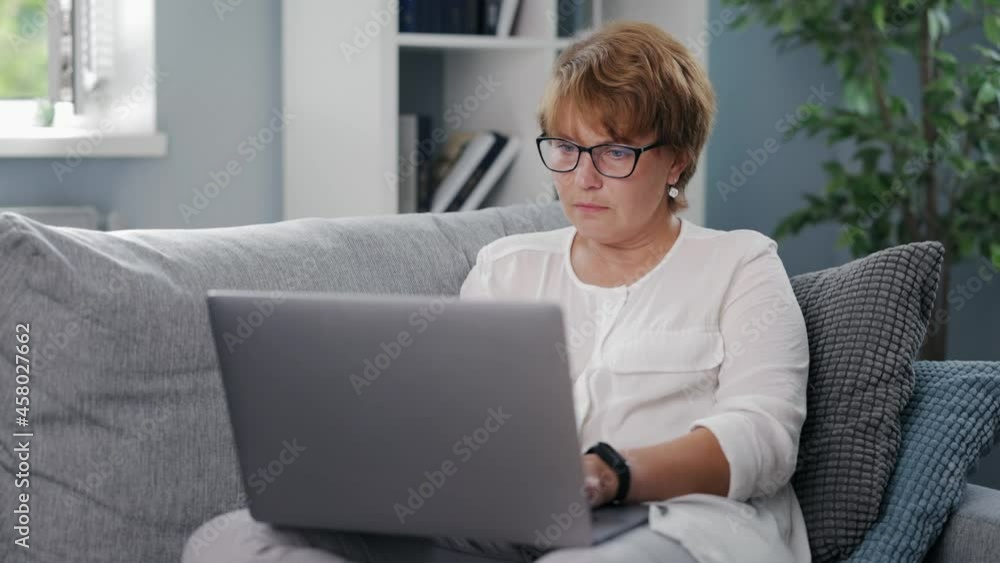 Focused senior woman in casual wear and eyeglasses sitting on sofa and working on wireless laptop. Concept of people, remote work and technology. Focused senior woman in casual wear and eyeglasses sitting on sofa and working on wireless laptop. Concept of people, remote work and technology.