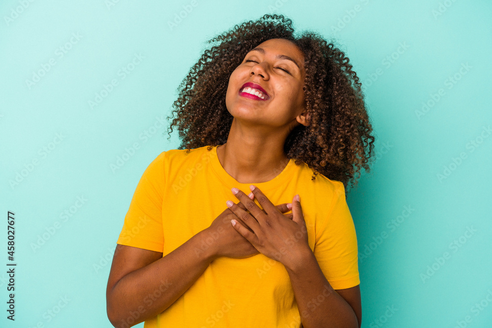 Young african american woman with curly hair isolated on blue background laughing keeping hands on heart, concept of happiness. - obrazy, fototapety, plakaty 
