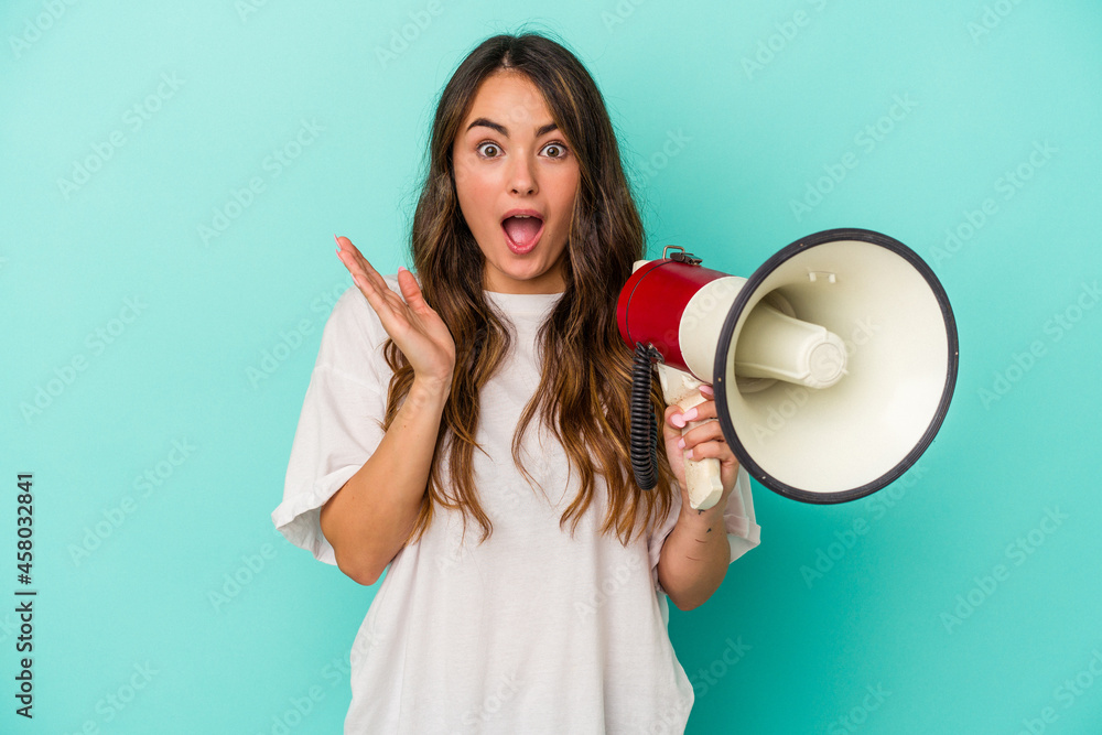 Young caucasian woman holding a megaphone isolated on blue background surprised and shocked. Young caucasian woman holding a megaphone isolated on blue background surprised and shocked.