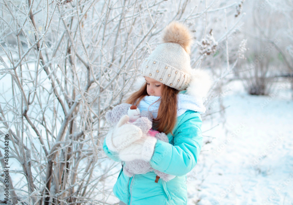 A small, charming girl in winter in a snowy park holds a teddy bear. Background of a winter forest landscape A small, charming girl in winter in a snowy park holds a teddy bear. Background of a winter forest landscape