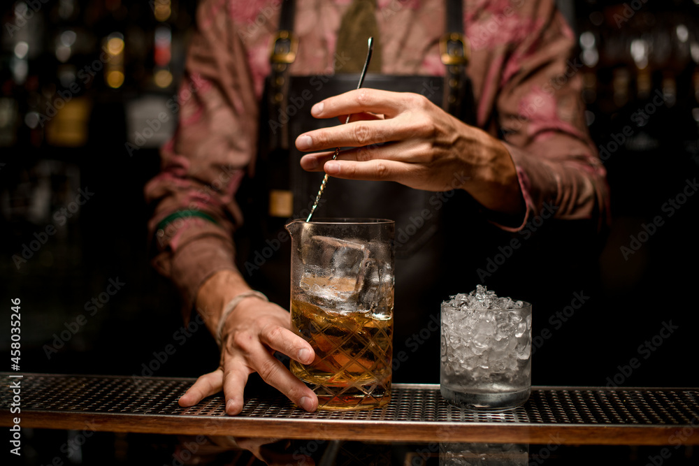 glass with crushed ice and mixing cup with liquid on the bar counter and hand of bartender stirring cocktail