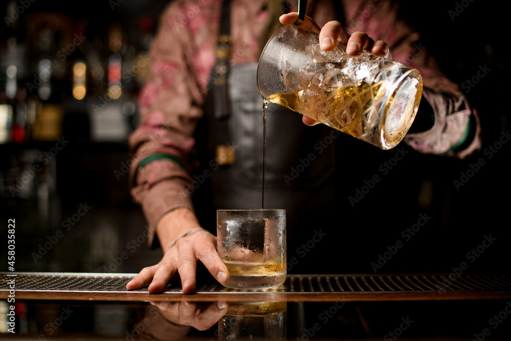 hand of barman holds crystal mixing cup with drink and pour it into a glass at the bar