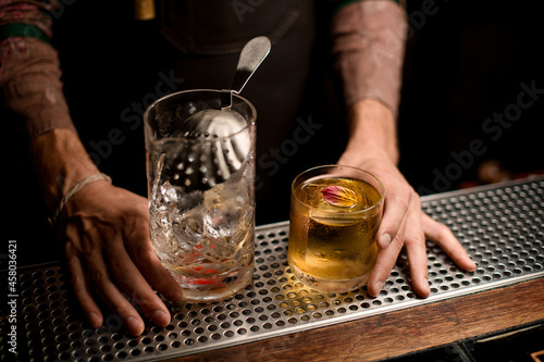 angle view of glassy mixing cup with ice and strainer on bar counter and glass with cocktail.