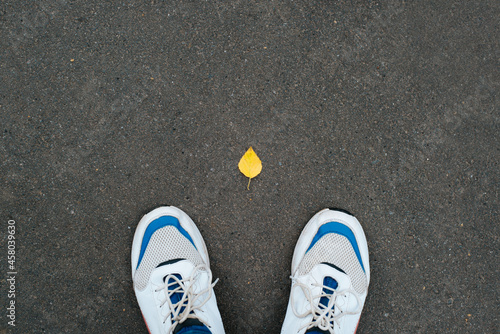 Man in shoes stands on asphalt with single fallen autumn yellow leaf, top view. Concept hello october, autumn season